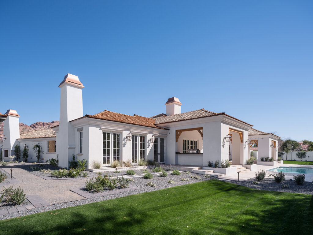 Rear exterior of a white stucco Mediterranean-style home with clay tile roofing, tall white chimneys with copper caps and a covered outdoor kitchen patio with timber brackets, overlooking gravel garden beds, a green lawn and the pool patio with distant mountain peaks