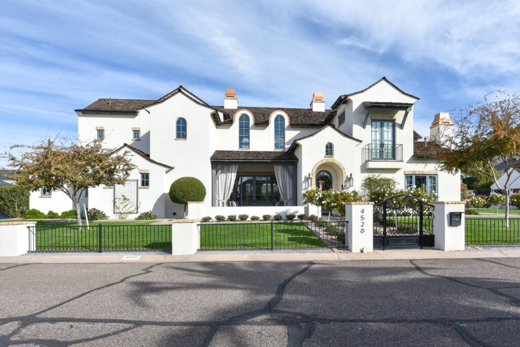 Front exterior of a two-story white stucco Spanish-Mediterranean home with dark shake roofing, terracotta chimney caps and dark-framed arched windows, behind a black wrought-iron fence with white masonry pillars on a manicured lawn